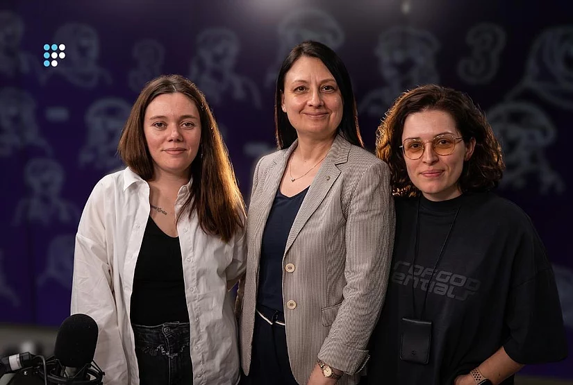 На фото - три жінки, які стоять разом перед камерою. Ліворуч-жінка у білій сорочці поверх чорного топа. У центрі-жінка у світлому смугастому піджаку. Праворуч-жінка в чорному одязі та окулярах. Унизу ліворуч видно студійний мікрофон. Фото зроблене під час запису у радіостудії.