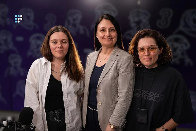 На фото - три жінки, які стоять разом перед камерою. Ліворуч-жінка у білій сорочці поверх чорного топа. У центрі-жінка у світлому смугастому піджаку. Праворуч-жінка в чорному одязі та окулярах. Унизу ліворуч видно студійний мікрофон. Фото зроблене під час запису у радіостудії.