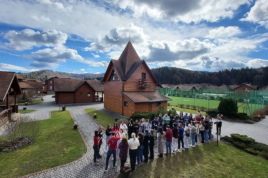 The photo shows a hotel complex with wooden houses. In the foreground, a group of 30-40 people gathered in a circle. It looks like they are taking part in a team building activity.