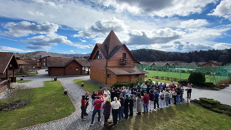 The photo shows a hotel complex with wooden houses. In the foreground, a group of 30-40 people gathered in a circle. It looks like they are taking part in a team building activity.